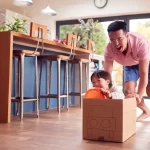 A dad pushing a boy in a cardboard box in his dining area.