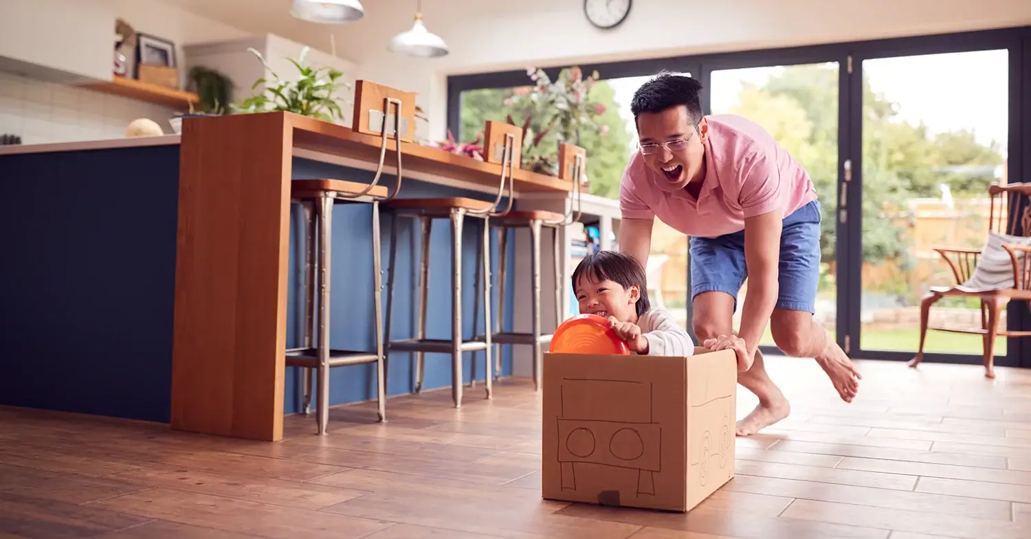A dad pushing a boy in a cardboard box in his dining area.