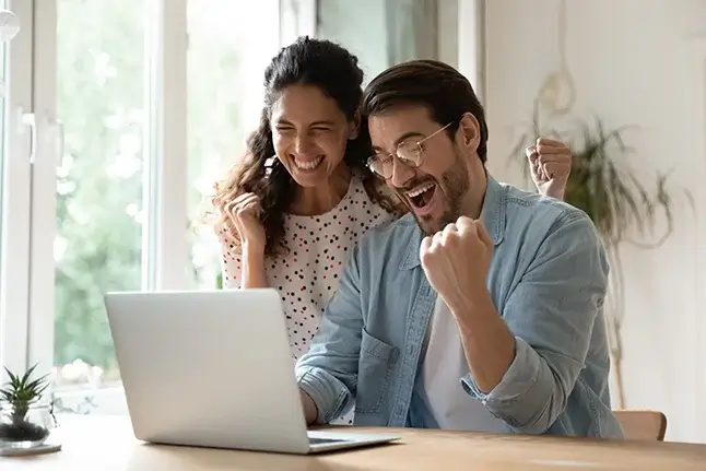 A couple raises their fists in triumph over something they accomplished on a laptop.