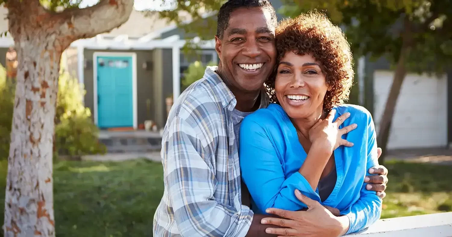 An image of a couple holding each other while posing along a fence in front of a house with a teal door.