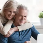 A older man looking at a computer while his partner hugs him from behind.